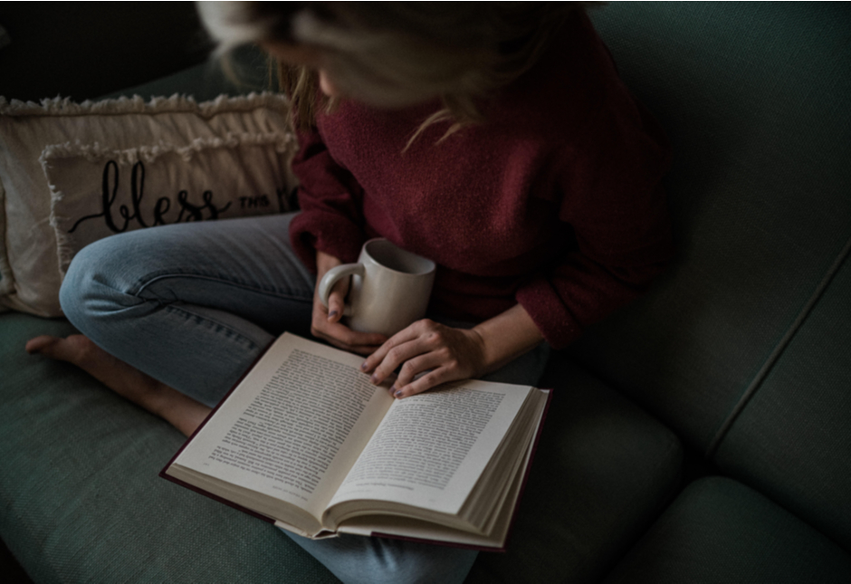 A woman sitting on a sofa reading a book with a cup of coffee