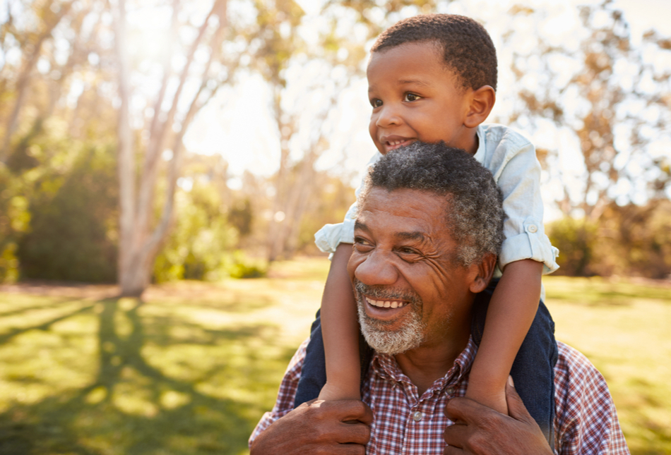 A grandfather carrying his grandson on his shoulders through a park.