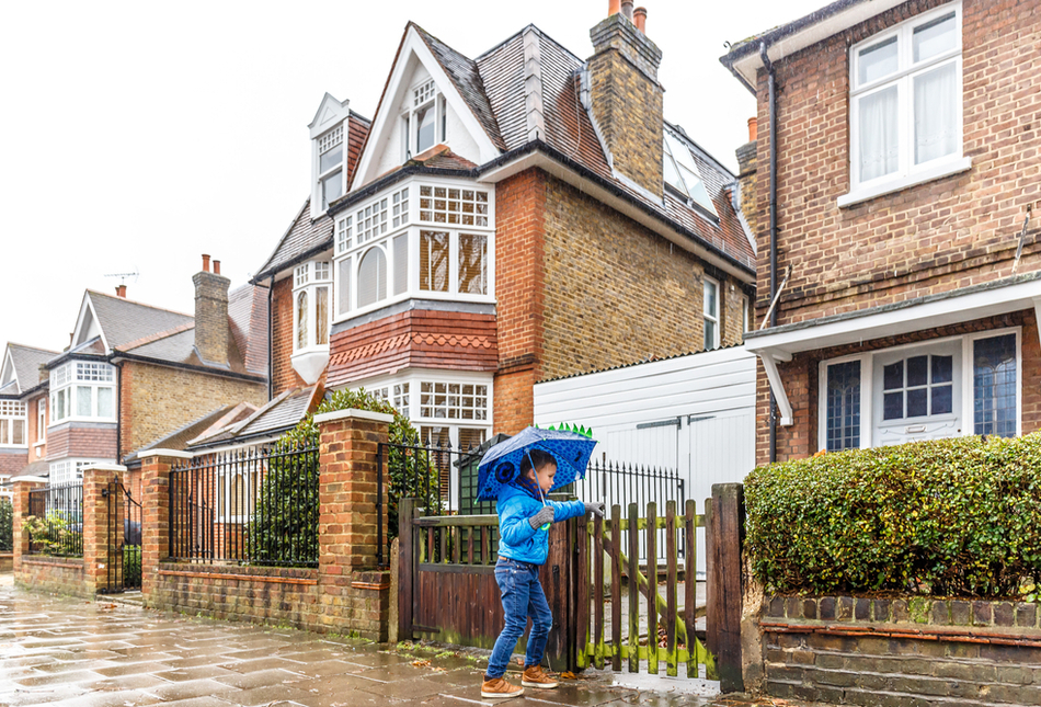 A boy with an umbrella opening a gate to a home.