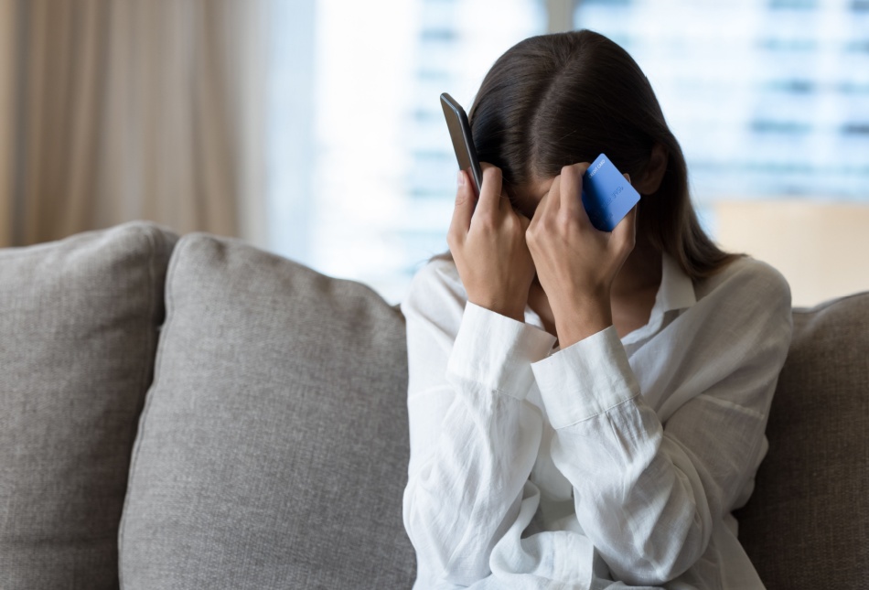 A woman holds her head with a phone in one hand and a bank card in the other.
